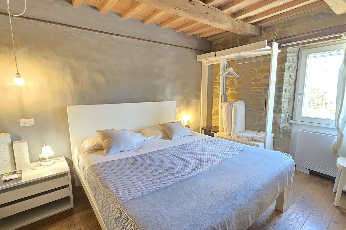 Una camera da letto con un letto grande, comodini, una finestra con tende, un muro in pietra e un soffitto in legno.