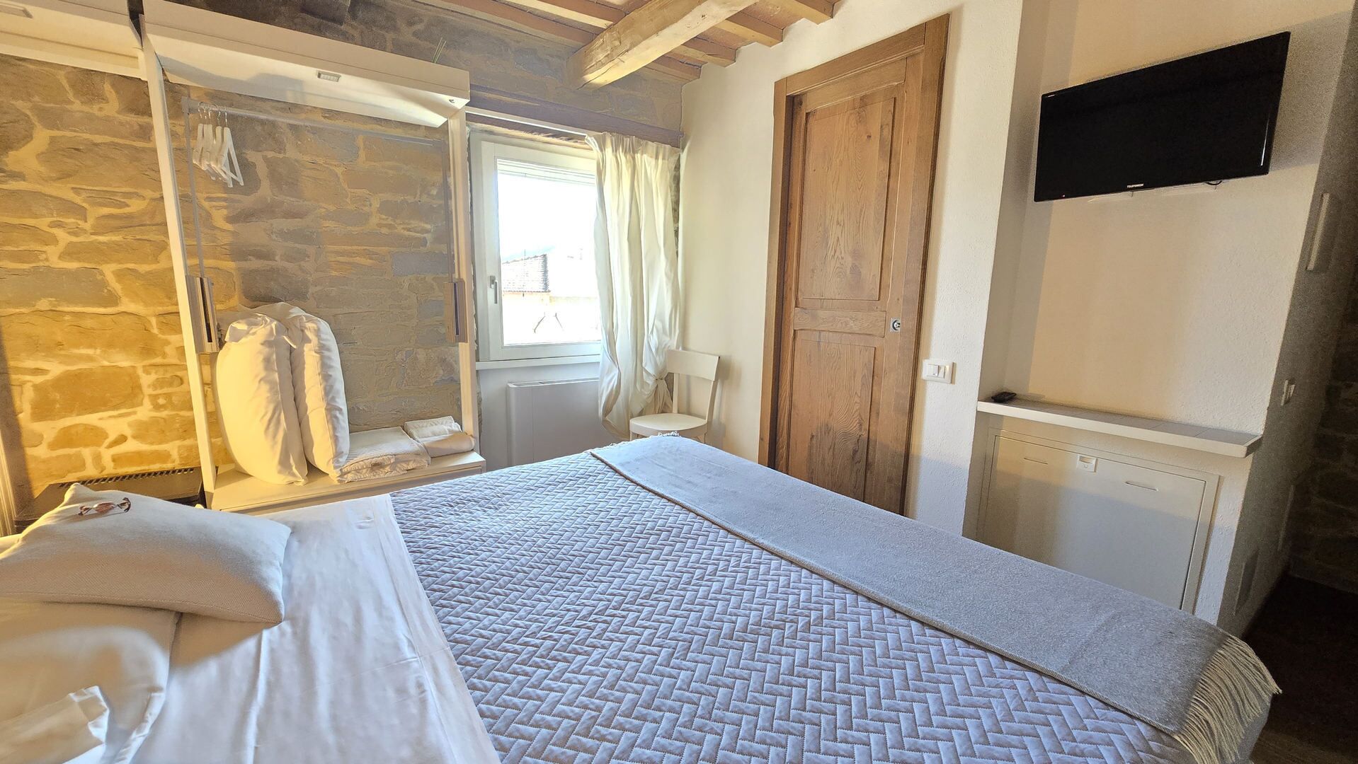 A bedroom with a bed, a stone wall, a wooden door, a TV, and a window with curtains.