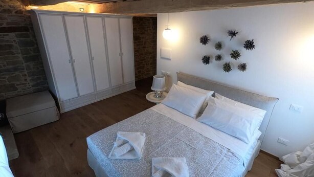 A bedroom with a bed, white curtains, a stone wall, and a ceiling with exposed beams. - Cà Maina (Santa Sofia)