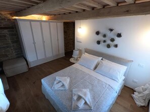 A bedroom with a bed, white curtains, a stone wall, and a ceiling with exposed beams. - Cà Maina (Santa Sofia)