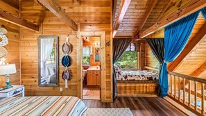 Ein Holzblockhaus-Innenraum mit einem Bett, einer kleinen Sitzecke, einem Spiegel und Blick ins Freie.