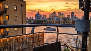 A balcony with wicker furniture, a telescope, and a view of a city skyline at sunset. - 2BR w/ Private Balcony & River Thames Views (London)