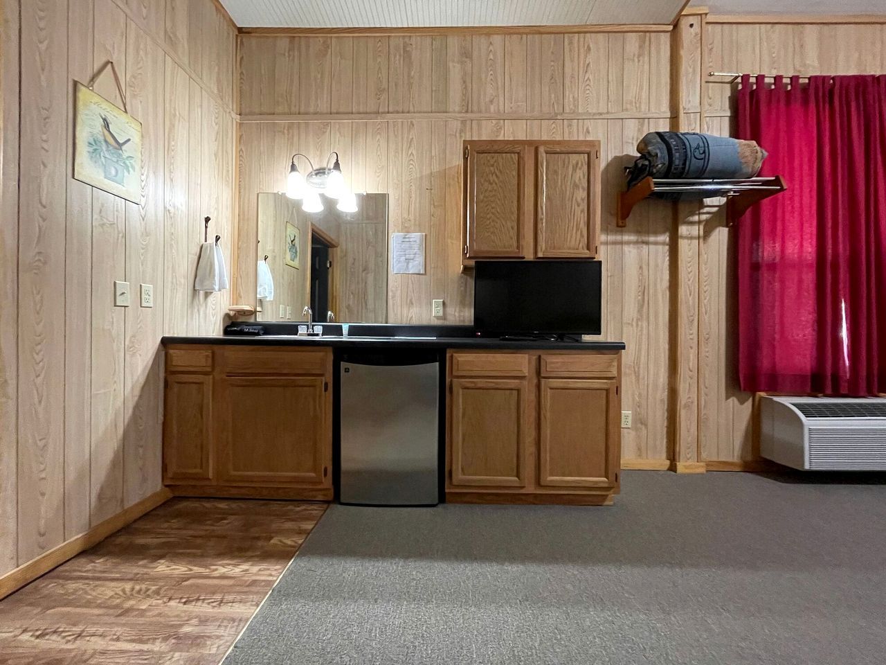 A compact kitchen with wooden cabinets, a built-in dishwasher, a flat-screen TV, and a red curtain.