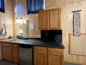 Une cuisine avec des meubles de rangement en bois, un lave-vaisselle, un plan de travail noir et un téléviseur fixé au mur.