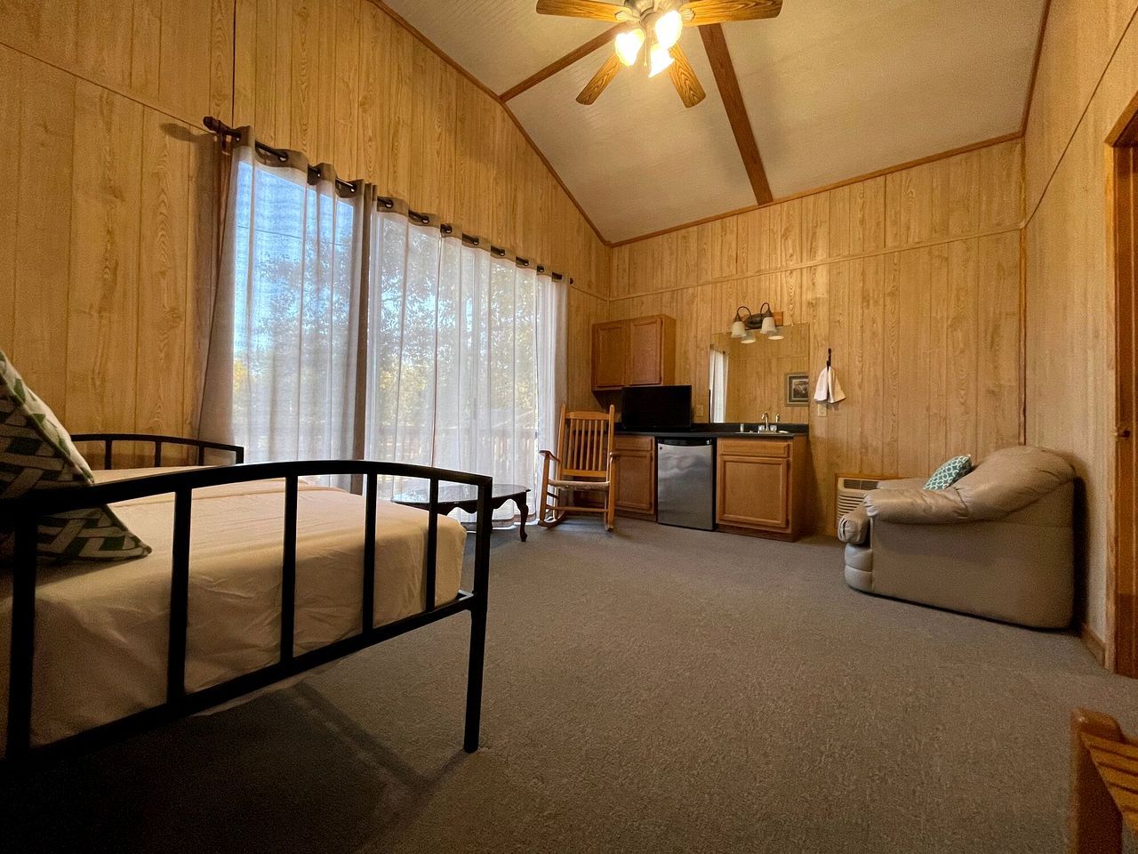 A room with a bed, a chair, a sofa, a kitchenette, and a ceiling fan.