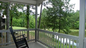 A covered porch with a rocking chair, white railing, and a view of a pond surrounded by trees.