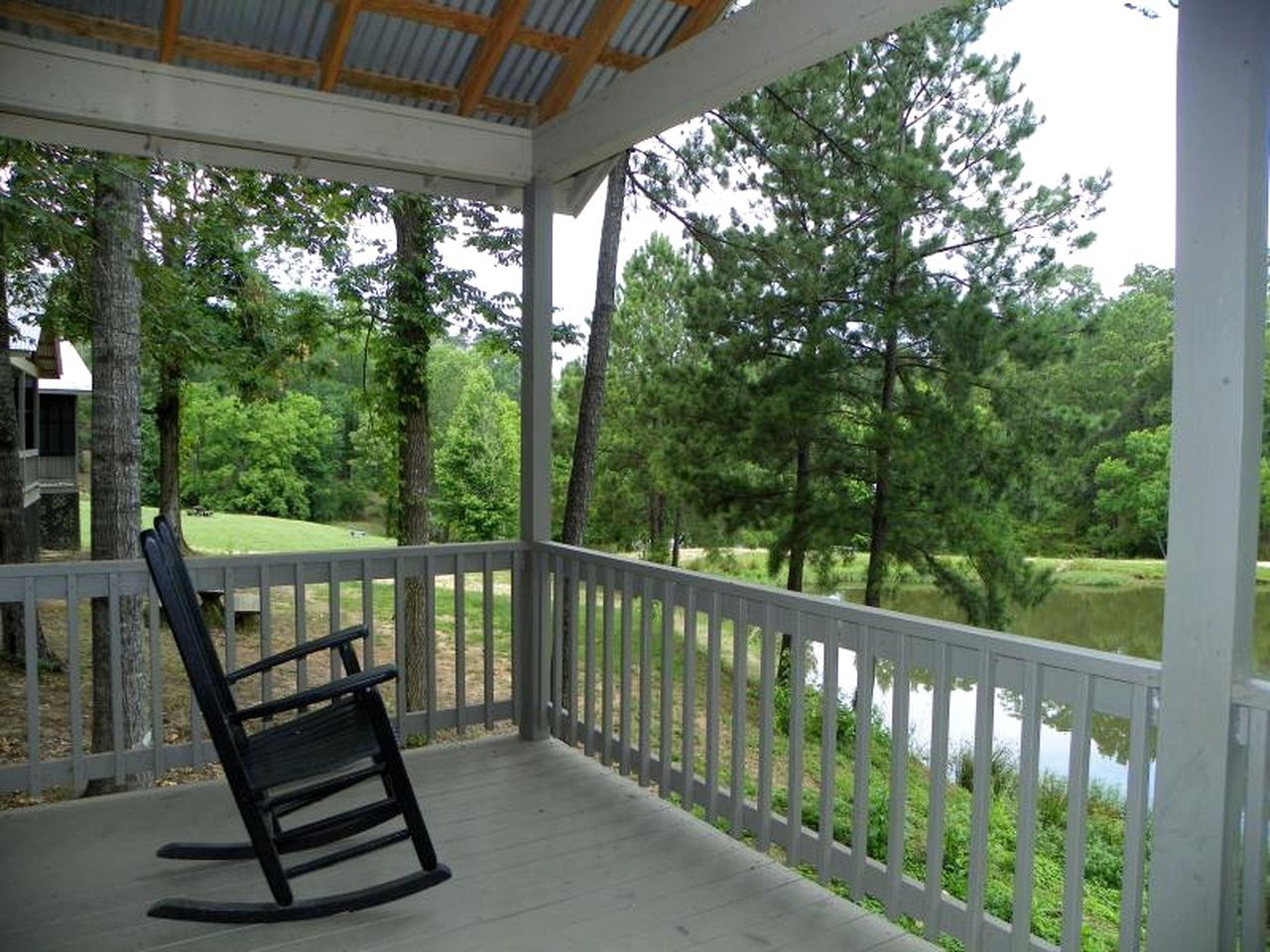 A covered porch with a rocking chair, white railing, and a view of a pond surrounded by trees.