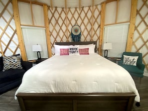 A bed with a white duvet, two pillows with text, a headboard, a wall with a lattice pattern, a window with blinds, a lamp, a chair with a cushion, and a decorative item on the wall.