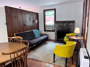 Un salon chaleureux avec un mur lambrissé, un canapé, une table à manger avec des chaises, une cheminée et une fenêtre donnant sur un beau paysage.