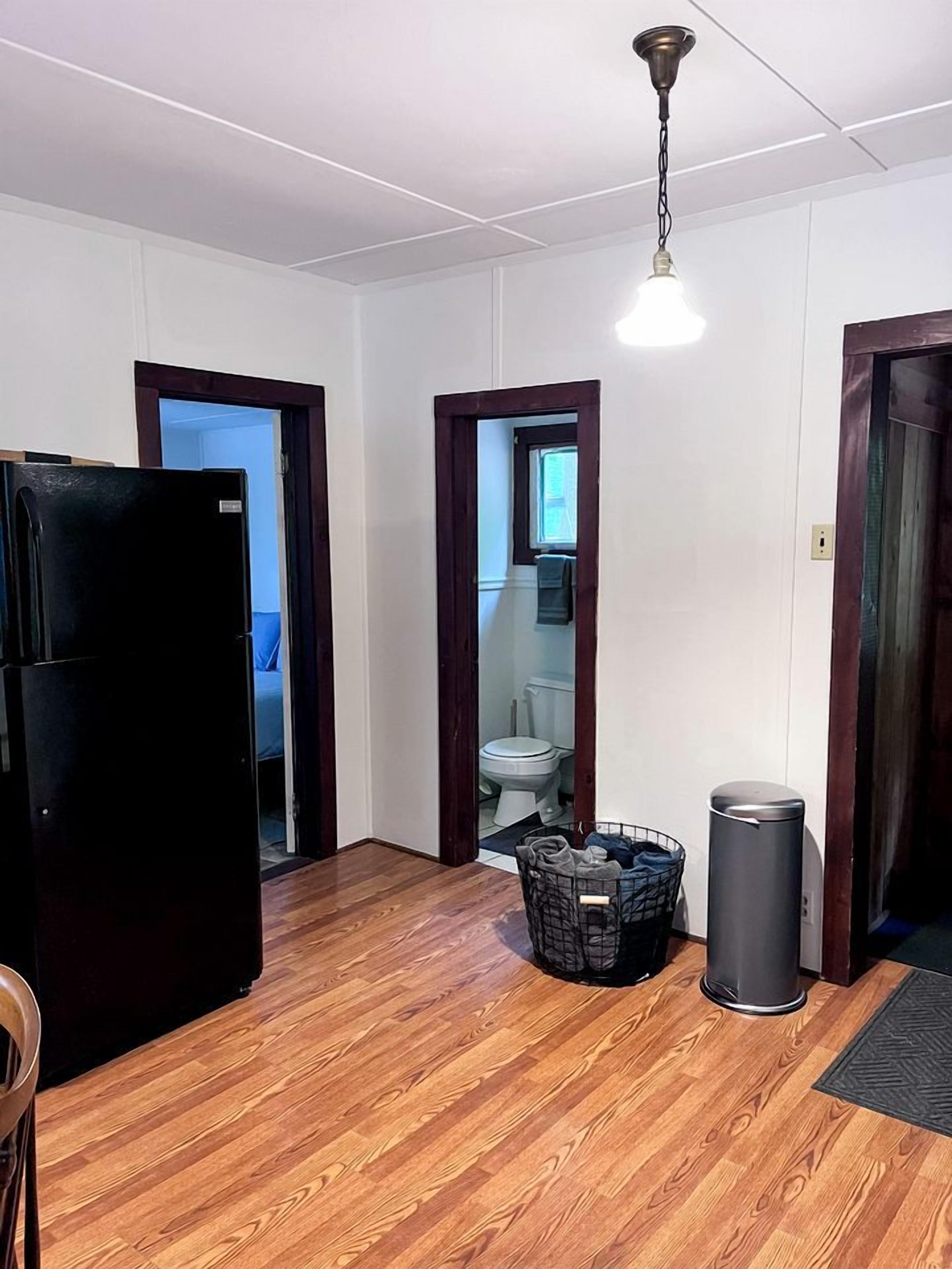 A room with wooden flooring, a black refrigerator, a laundry basket, a trash can, and a hanging light fixture.
