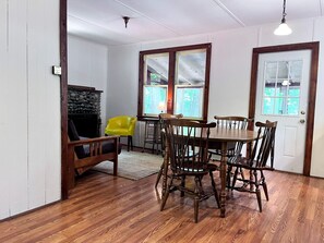 A dining area with a wooden table and chairs, a yellow chair, and a fireplace. - Charming One-Floor Waterfront Cabin with Dock & Fire Pit Near the River | Corinth, New York (Corinto)