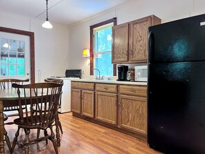 A kitchen with wooden cabinets, a black refrigerator, a microwave, and a dining table with chairs. - Charming One-Floor Waterfront Cabin with Dock & Fire Pit Near the River | Corinth, New York (Corinto)