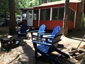 Una cabaña rústica con fachada roja, un porche con sillas Adirondack azules y una fogata.