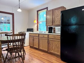 A kitchen with wooden cabinets, a black refrigerator, a microwave, and a dining table with chairs. - Charming One-Floor Waterfront Cabin with Dock & Fire Pit Near the River | Corinth, New York (Corinto)