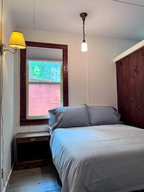 A bedroom with a bed, a window with blinds, a nightstand, and a lamp. - Charming One-Floor Waterfront Cabin with Dock & Fire Pit Near the River | Corinth, New York (Corinto)
