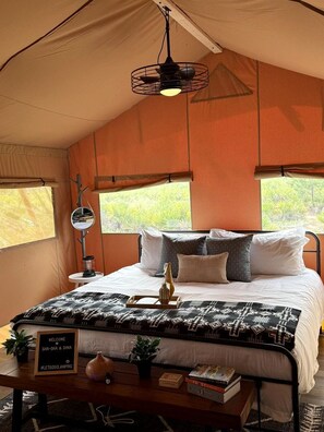 A tent with a bed, a hanging light fixture, a small table with a sign, and a view of greenery outside.