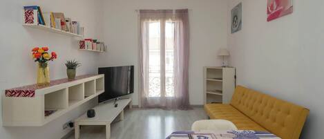 A living room with a sofa, TV, shelves, and a window with curtains.