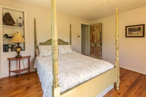 Een hemelbed met een bloemenlaken, een houten nachtkastje met een lamp, een boekenplank met boeken en decoratieve objecten, en een deur met een rustieke uitstraling.