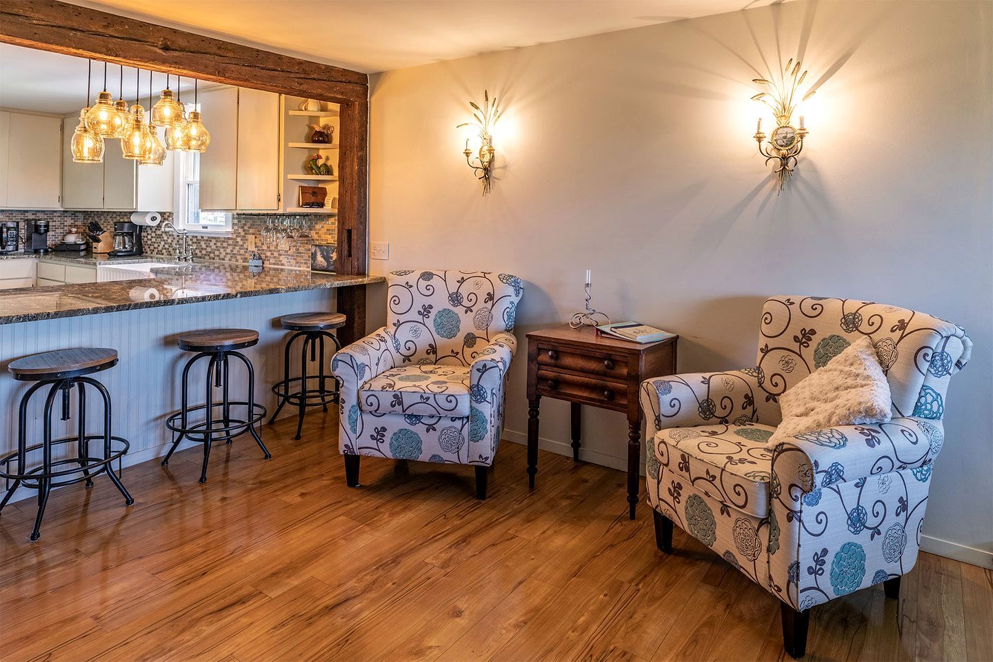 A kitchen with a bar area, two patterned armchairs, a small table, and a wall-mounted light fixture.