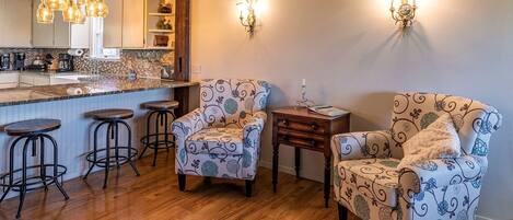 A kitchen with a bar area, two patterned armchairs, a small table, and a wall-mounted light fixture.
