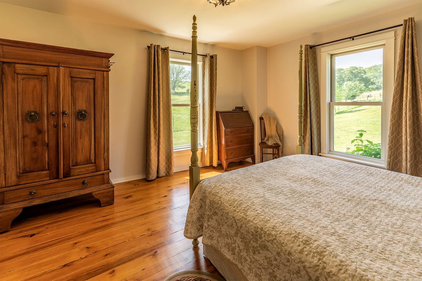 A bedroom with a wooden wardrobe, a four-poster bed, a wooden dresser, and a view of a green landscape through the window.