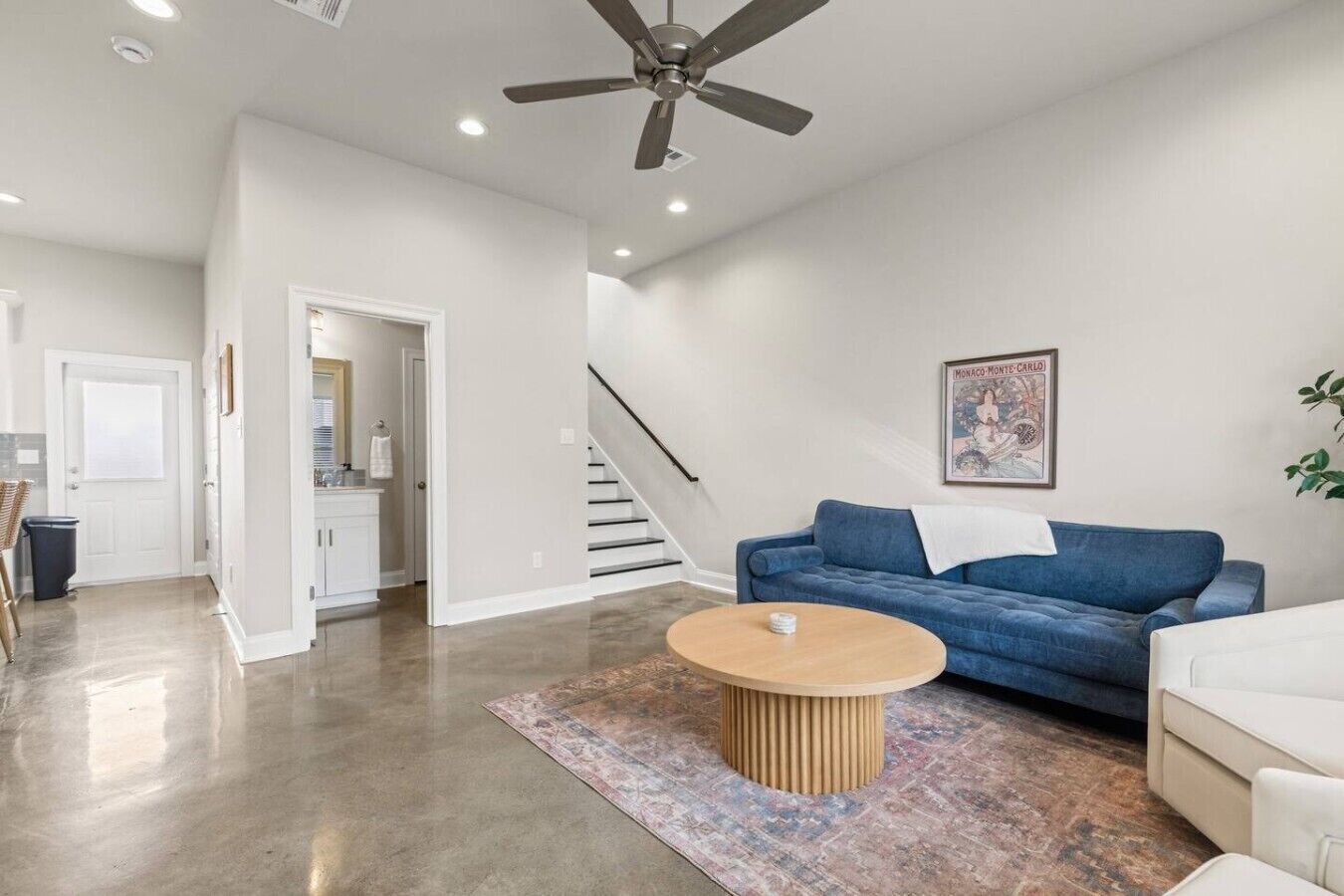 Un salon spacieux avec un canapé bleu, une table basse en bois et un ventilateur de plafond. Un escalier mène à l’étage supérieur, et un tableau encadré est accroché au mur.