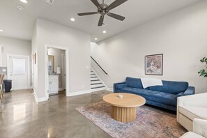 Amplia sala de estar con un sofá azul, una mesa de centro de madera y un ventilador de techo. Una escalera conduce a un nivel superior, y hay un cuadro enmarcado en la pared.