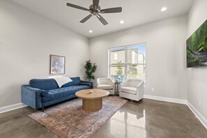 A living room with a blue sofa, a round coffee table, two white chairs, a ceiling fan, and a window with blinds.