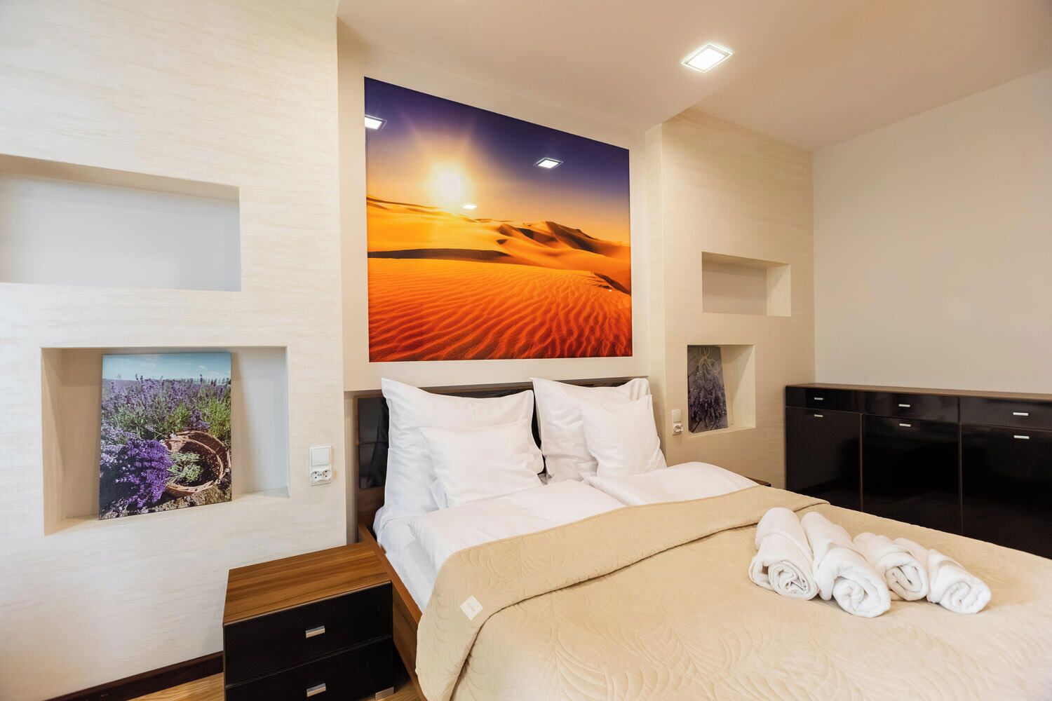 Ein Hotelzimmer mit einem Bett, einem großen Wandbild eines Wüstensonnenuntergangs, einem kleinen gerahmten Bild von Lavendel und einem ordentlich bezogenen Bett mit Handtüchern.