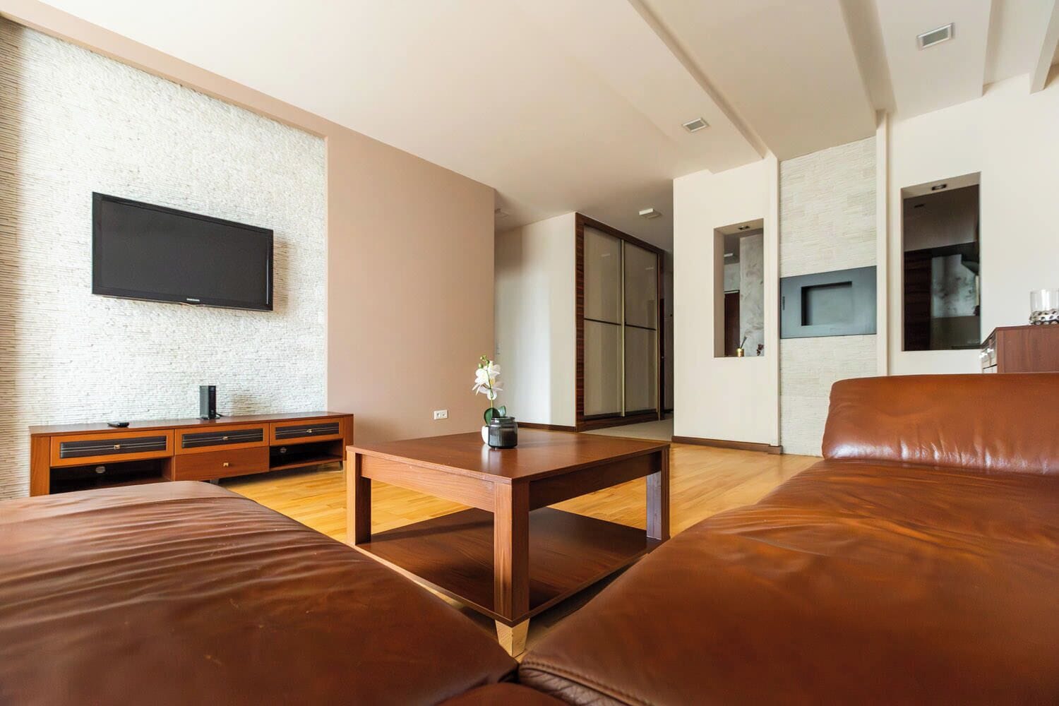 Een moderne woonkamer met een flatscreen televisie aan een gestructureerde muur, een houten salontafel en een bruine leren bank.