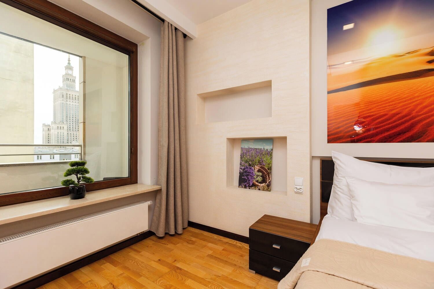 A hotel room with a large window, a small potted plant, a nightstand, a bed, and a wall-mounted artwork.