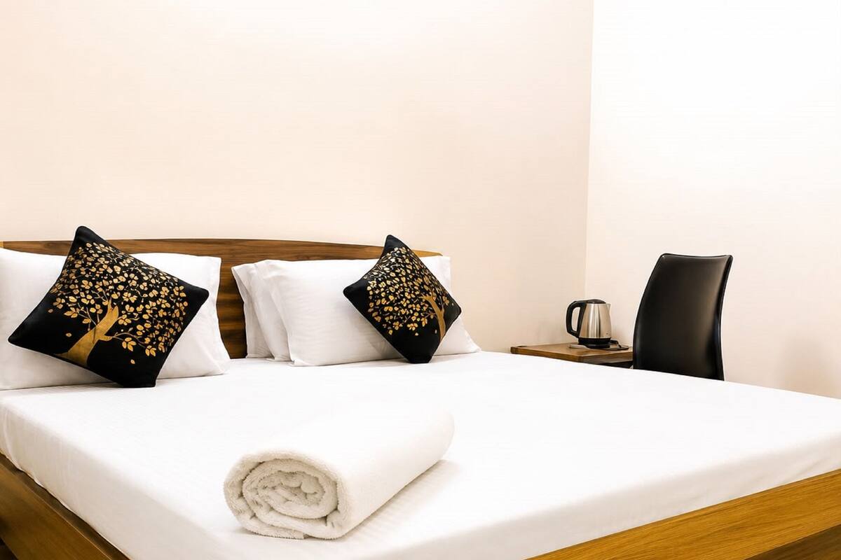 Ein ordentlich gemachtes Bett mit weißen Leinentüchern, zwei goldene und schwarze Zierkissen, ein aufgerolltes weißes Handtuch, ein Holz-Kopfteil, ein kleiner Nachttisch mit Wasserkocher und ein schwarzer Stuhl.