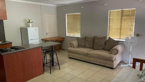 A living room with a sofa, a dining table, a kitchen area with a refrigerator, and a window with blinds.