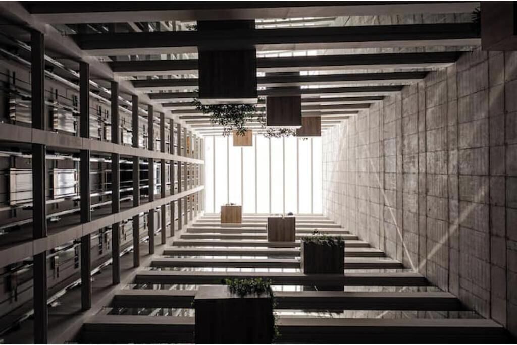 A modern library interior with bookshelves, a central staircase, and hanging plants.
