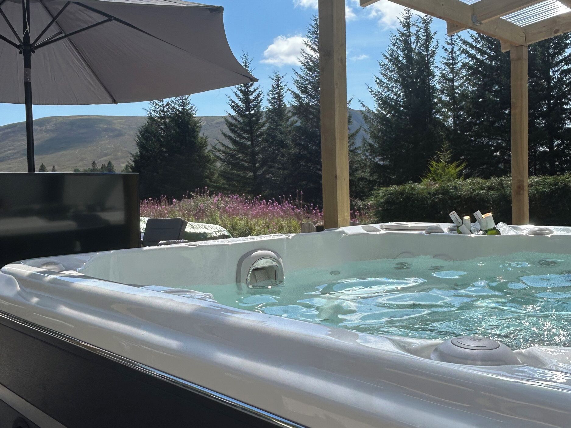 Ein Whirlpool mit einer hölzernen Pergola darüber, umgeben von Bäumen und Blick auf die Berge.