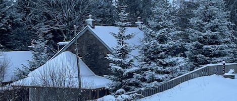 A snow-covered house with a stone exterior, surrounded by trees and a wooden fence.
