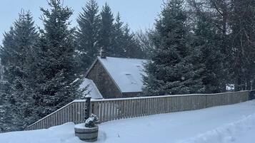 A snow-covered landscape with a wooden fence, a small tree in a barrel planter, and a house with a sloped roof.