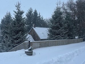 A snow-covered landscape with a wooden fence, a small tree in a barrel planter, and a house with a sloped roof. - 1 Station Cottages (Pitlochry)