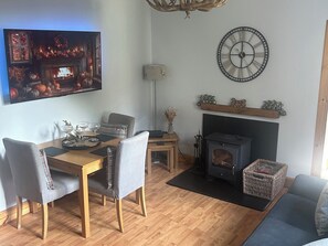 A cozy living room with a fireplace, a dining table with chairs, a TV mounted on the wall, and a clock on the wall. - 1 Station Cottages (Pitlochry)