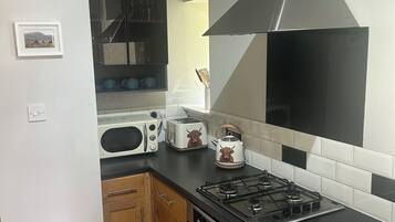 A kitchen with a gas stove, a microwave, a toaster, and a kettle. There is a wooden cabinet and a white wall with a picture frame.