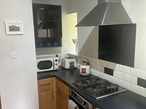 Una cocina con estufa a gas, microondas, tostadora y hervidor. Hay un armario de madera y una pared blanca con un marco de fotos.