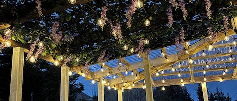Una pérgola de madera con luces de cuerda y flores moradas.