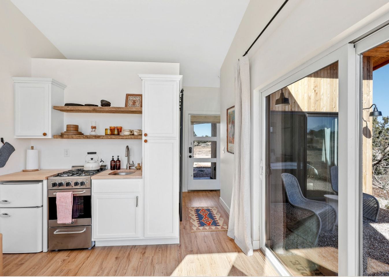 Una cucina compatta con mobili bianchi, un fornello e un lavello. C'è una zona pranzo con tavolo e sedie e un'ampia porta scorrevole in vetro che dà su uno spazio esterno.
