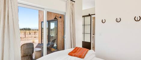 A bedroom with a large bed, a view of the desert through a sliding glass door, a small table with a chair, and a glass door leading to a balcony.