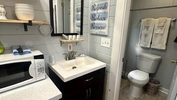 A compact bathroom with a white sink, a microwave, a mirror, and a toilet. There are signs on the wall that read 'Refresh Relax' and 'Renew.'