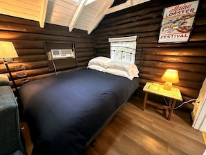 Ein Blockhaus-Zimmer mit einem Bett, einem kleinen Tisch mit Lampe, einem Fenster und einem Poster an der Wand.