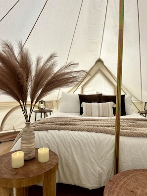 Eine Zelteinrichtung mit einem Bett, einer Vase mit Trockenpflanzen und zwei brennenden Kerzen auf einem Holztisch.