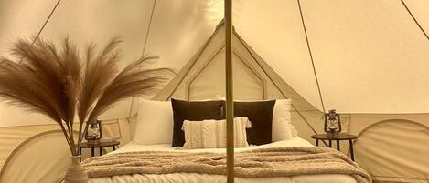 A tent with a bed, two wooden side tables, a hanging light fixture, and a vase with dried plants.