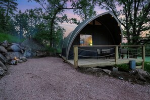 Eine halbkreisförmige, überdachte Außenanlage mit Holzterrasse und Sitzgelegenheit, umgeben von einer natürlichen Landschaft und einem Kiesweg.