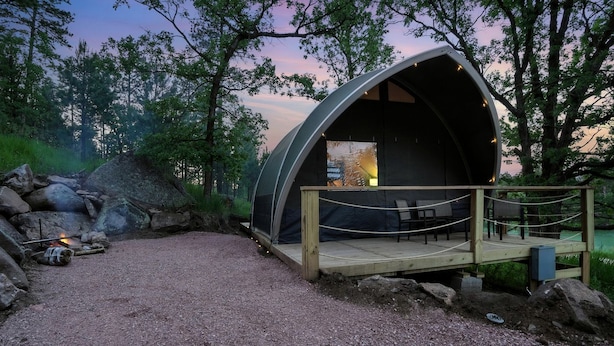 Une structure extérieure semi-circulaire et abritée, dotée d’une terrasse en bois et d’un espace salon, entourée d’un paysage naturel et bordée d’un chemin de gravier.